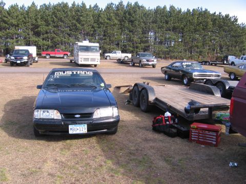 1989 Ford Mustang - A black 1989 Ford Mustang is parked at a race track with other vehicles and trailers.