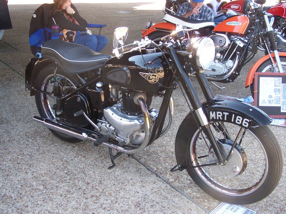 Black BSA motorcycle with license plate MRT 186, parked near an orange motorcycle and people in chairs