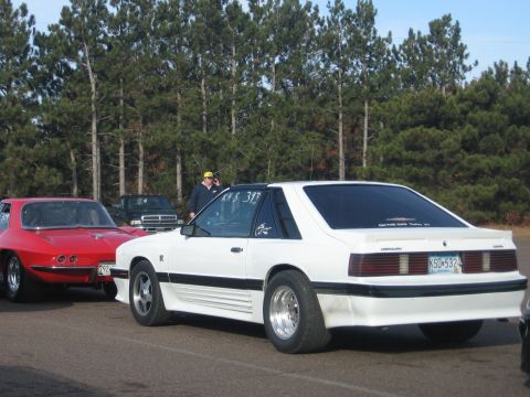 1989 Ford Mustang - A white Ford Mustang, likely from the late 80s or early 90s, is parked at a race track. A red Chevrolet Corvette is to its left.