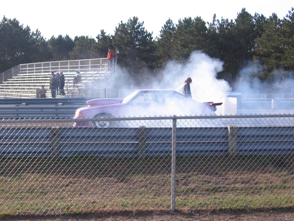 Drag car doing a tire-smoking burnout behind a fence at a track