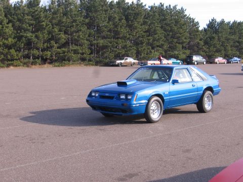1989 Ford Mustang - Blue coupe with a prominent hood scoop and drag racing tires sits on asphalt with trees in the background.