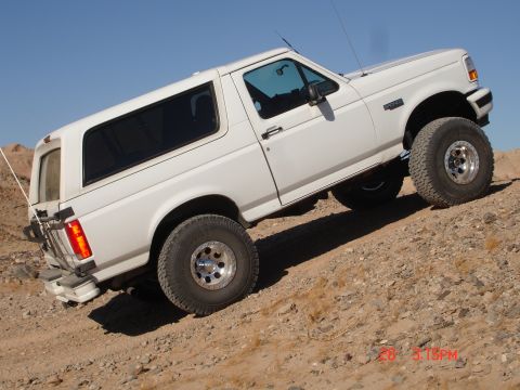 1996 Ford Bronco - White 1996 Ford Bronco is shown off-road on a sandy incline under a clear blue sky.