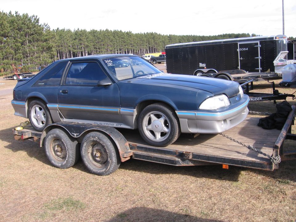 Dark blue coupe on a trailer, with blue and silver trim. "Mustang GT" visible on the side