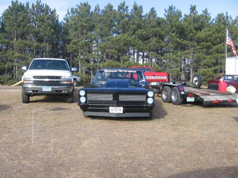 1989 Ford Mustang - A black drag car with a prominent hood scoop and custom grille sits parked at a race track, with other vehicles and a trailer nearby.