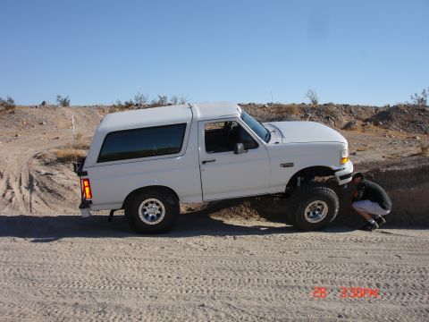 1996 Ford Bronco - A white 1996 Ford Bronco is shown in a desert setting with a man crouching to inspect the front wheel.