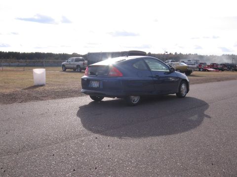 1989 Ford Mustang - A blue coupe is parked on a track with other vehicles and trailers in the background under a cloudy sky.