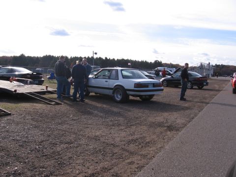 1989 Ford Mustang - Several people stand around a white coupe with wide rear tires at an outdoor event.
