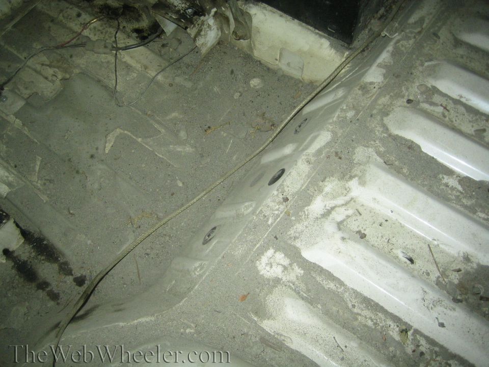 Close-up of a dirty, dusty vehicle floor pan with visible wiring and some white painted metal sections