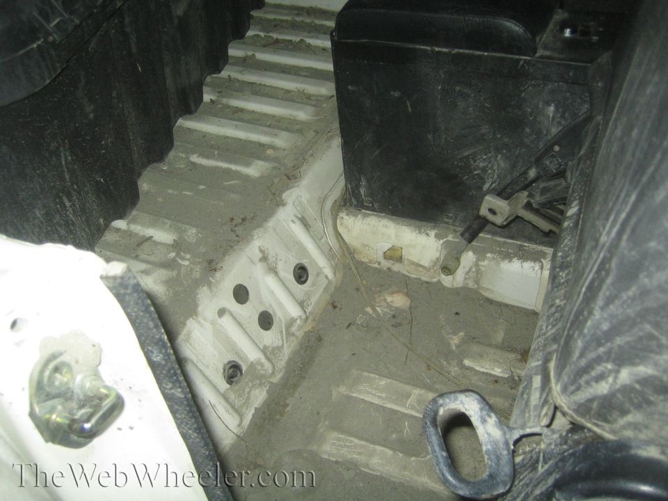 Interior floor of a vehicle showing the battery and some mounting hardware. The floor has a ribbed texture and appears dirty