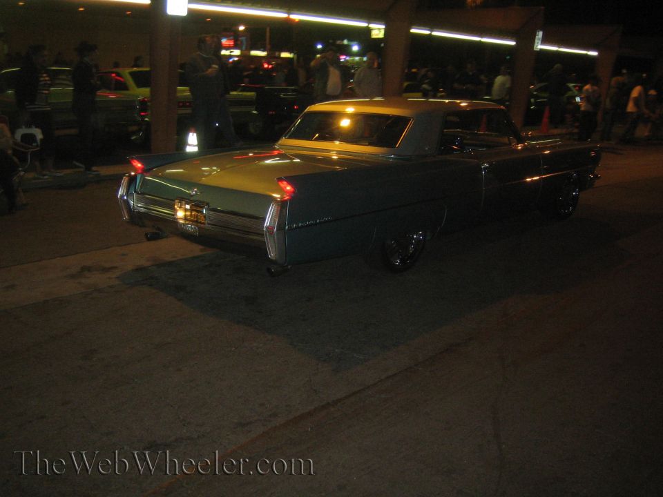 Rear view of a light blue Cadillac Coupe de Ville with a white vinyl top. It has prominent tail fins and chrome trim. Other cars and people are visible in the background at night