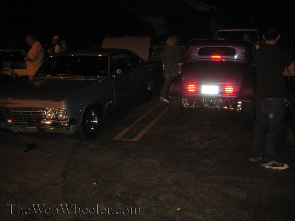 Two classic cars parked side-by-side at night. The car on the left is a light blue 1965 Chevrolet Impala. The car on the right is a dark purple hot rod