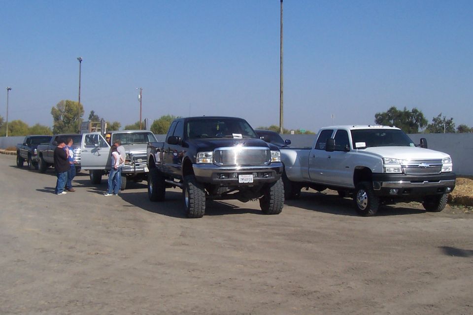 Several pickup trucks are parked in a lot under a clear blue sky. A dark blue truck is in the foreground, with a white dually truck to its right. Other vehicles and people are visible in the background