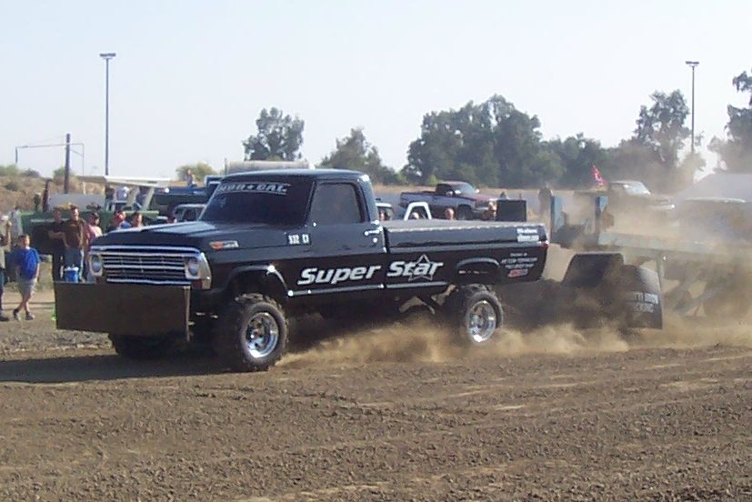 Black Ford pickup truck with 'Super Star' on the side, pulling a sled in a dirt arena. Other vehicles and people in background