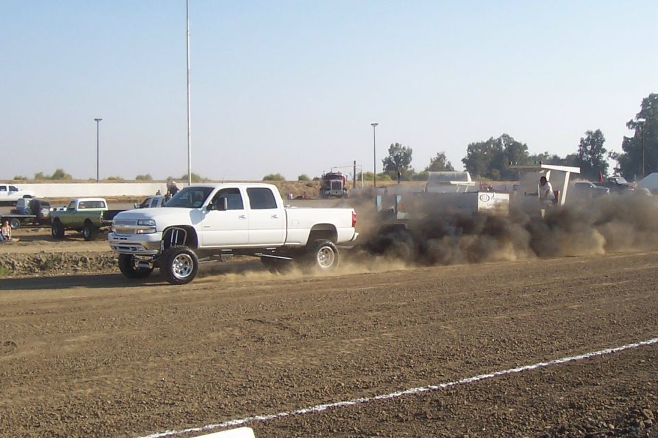 White pickup truck with lifted suspension is pulling a sled, kicking up a lot of black smoke. Another green truck is visible in the background