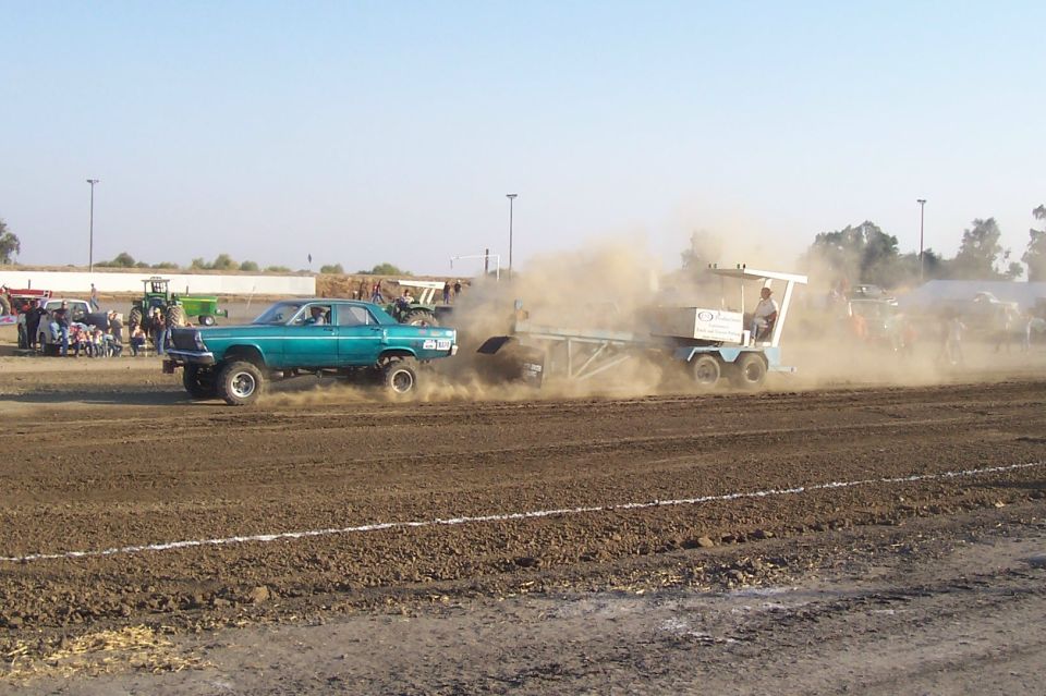 Modified teal sedan with large tires pulling a sled in a dirt arena, with tractors and spectators in the background