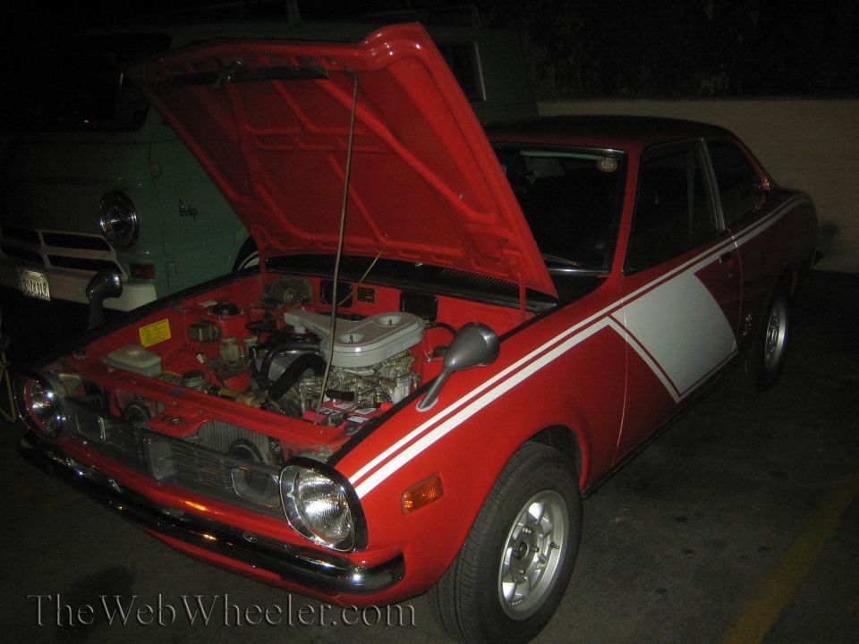 Red coupe with white stripes, hood open showing engine. Another vehicle is partially visible behind it