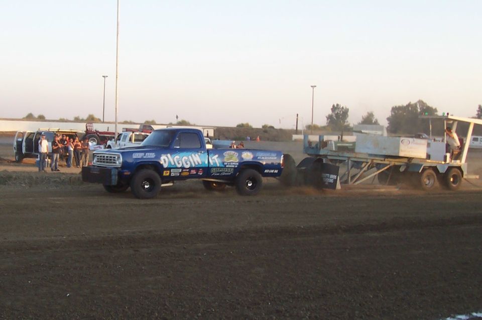 Blue pickup truck with 'DIGGIN IT' graphics pulling a sled on a dirt track. Other vehicles and people are visible in the background