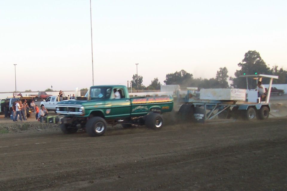 Green pickup truck with lifted suspension and large tires, towing a trailer with a person on it