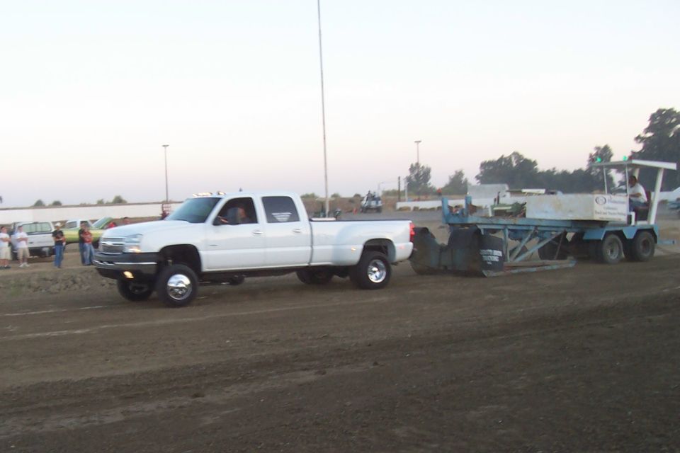 White dually pickup truck with chrome wheels is pulling a sled on a dirt track. Another vehicle is visible in the background