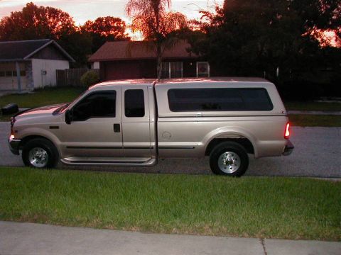 1999 Ford F-250 - A tan 1999 Ford F-250 Super Duty pickup truck with a camper shell is parked on the side of the road at dusk.