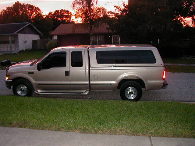 Tan Ford Super Duty extended-cab pickup with matching bed cap parked on a street at dusk