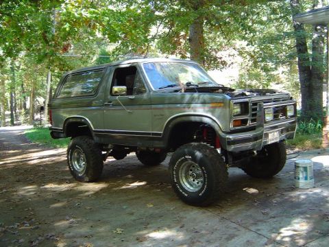 1986 Ford Bronco - A lifted green Ford Bronco with off-road tires sits on a dirt path surrounded by trees.
