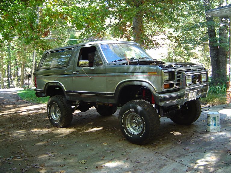 Green lifted Ford Bronco with off-road tires parked on a dirt path under trees