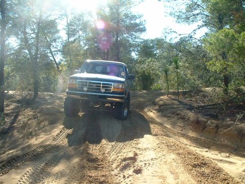 1995 Ford Bronco - A dark SUV navigates a sandy off-road trail under a bright, sunny sky.