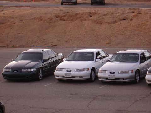 1993 Ford Taurus - Three Ford Taurus sedans are parked in a lot. The dark green car is on the left, followed by two white and silver models.
