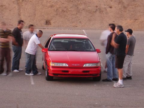 1993 Ford Taurus - A red coupe is surrounded by several people on a paved surface, possibly a parking lot.