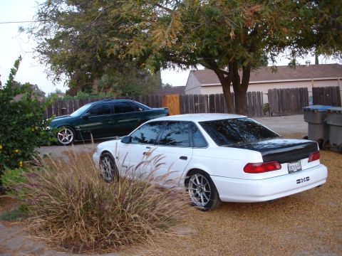 1993 Ford Taurus - A white sedan with a black trunk lid and aftermarket wheels is parked in a gravel driveway next to a green sedan.