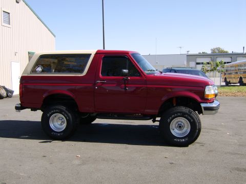 1989 Ford Bronco - Passenger-side profile of a lifted burgundy/red full-size Ford Bronco with a tan-topped removable hardtop, parked on asphalt beside a metal…