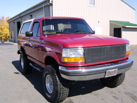 1989 Ford Bronco - Three-quarter front view of a glossy red two-door Ford Bronco with a tan removable hardtop, parked on asphalt outside a tan metal shop buil…