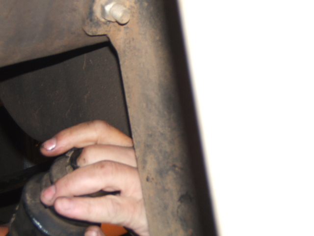 Hand reaching through access hole in rusty frame rail to grip round black part