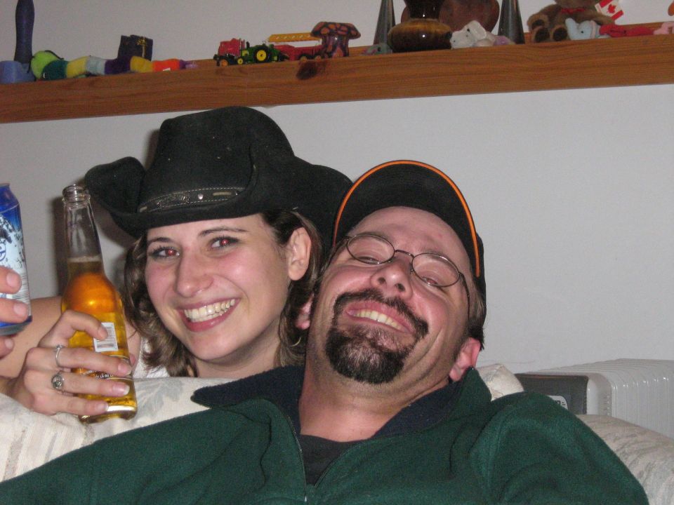 People smiling, one wearing a cowboy hat and holding a beer