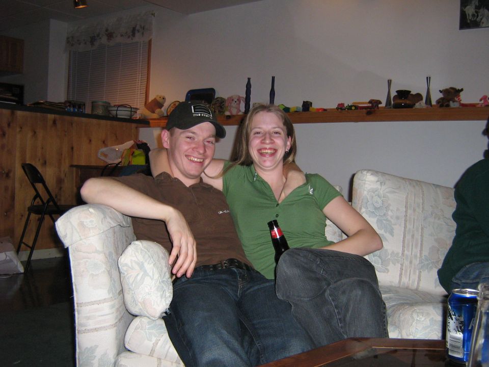 Guy in a baseball cap and a girl in a green shirt sit on a couch, both smiling. A beer bottle is on the couch