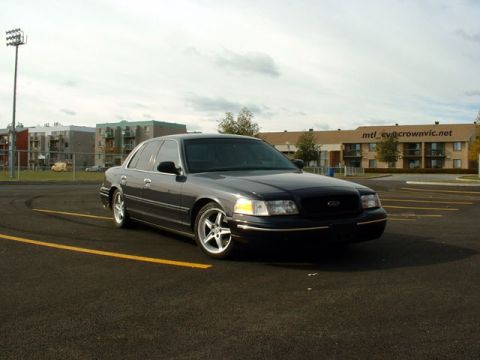 2000 Ford Crown Victoria - A dark-colored sedan is parked in an empty lot with buildings in the background.