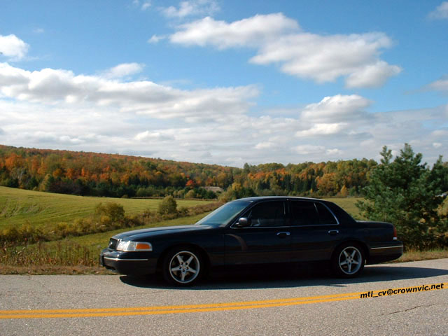 Black Ford Crown Victoria sedan side view parked along a rural road with fall foliage behind
