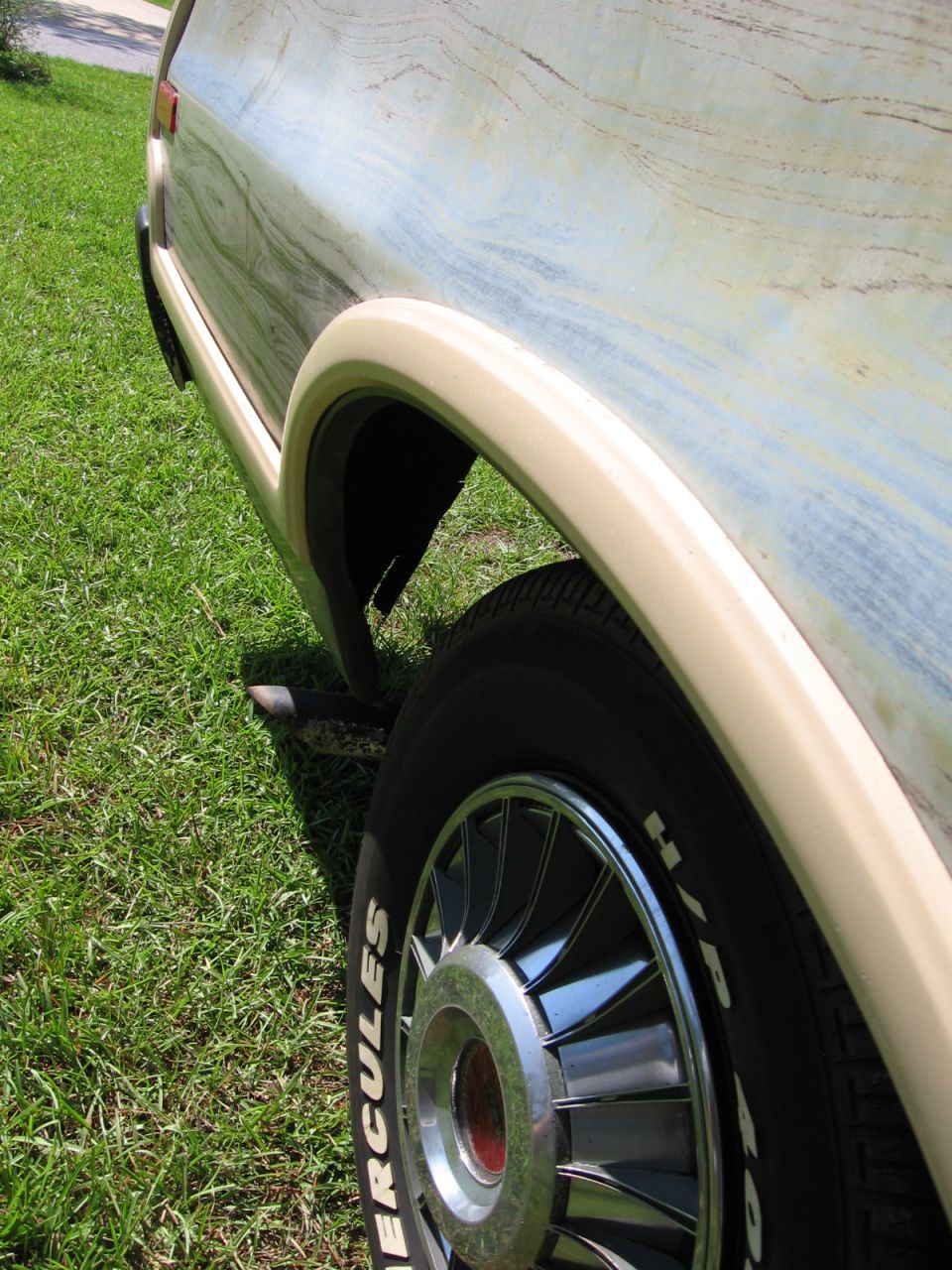 Vehicle's rear quarter panel with woodgrain-like paint and a beige fender flare over a wheel with a chrome hubcap and white-lettered tire
