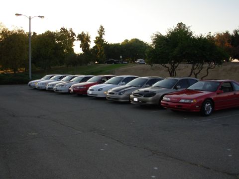 1995 Ford Taurus - A line of cars parked in a lot at dusk, with trees and a hill in the background.