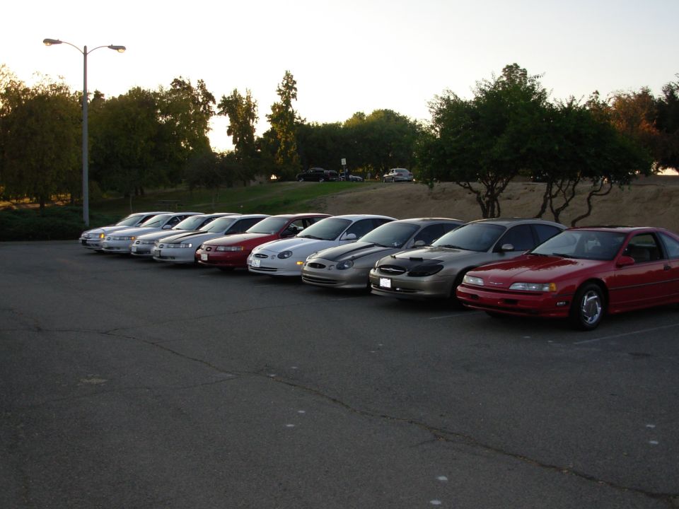 Several cars parked in a row, including a red coupe and a gold sedan with a black car cover