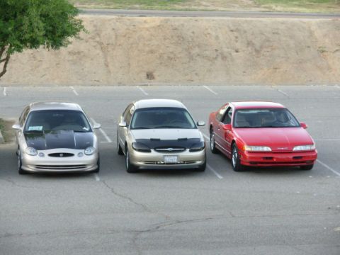 1995 Ford Taurus - Three cars are parked in a lot, with a tree on the left and a dirt embankment in the background.