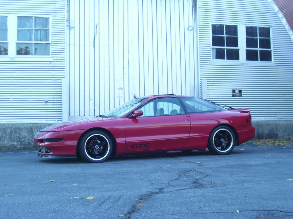 Red 1997 Ford Probe GTS coupe with black rims parked outside a corrugated metal building