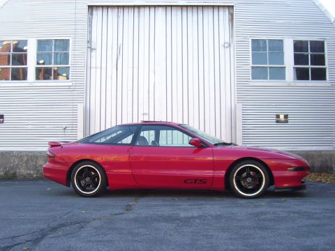1997 Ford Probe - A red coupe with a black roof is parked in front of a white building with windows.
