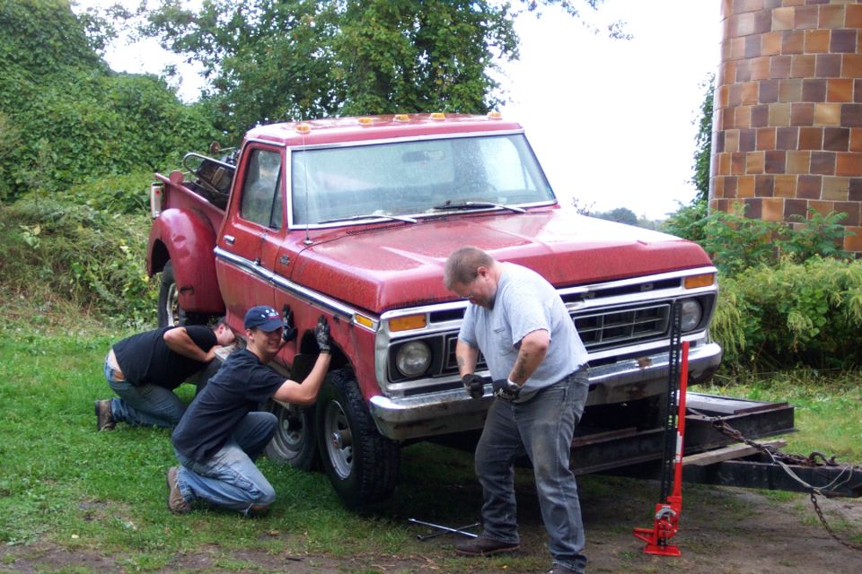 Red 1975 Ford F-150 pickup truck, front 3/4 view, with three men working on the front fender and wheel area