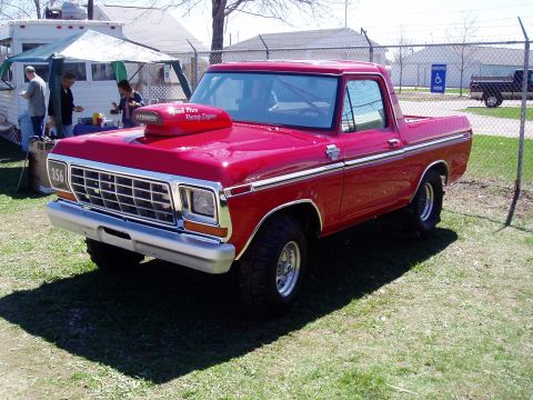 1986 Ford Bronco - A red Ford Bronco with a prominent hood scoop and racing numbers is parked on a grassy area near a trailer.