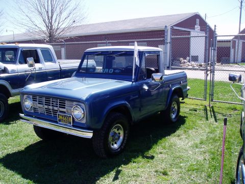 1986 Ford Bronco - A blue Ford Bronco is parked on a grassy area in front of a red building with a chain-link fence.
