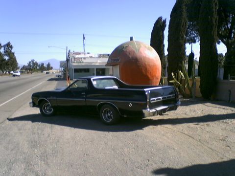 1973 Ford Ranchero - A black 1973 Ford Ranchero is parked next to a large roadside orange attraction on a sunny day.