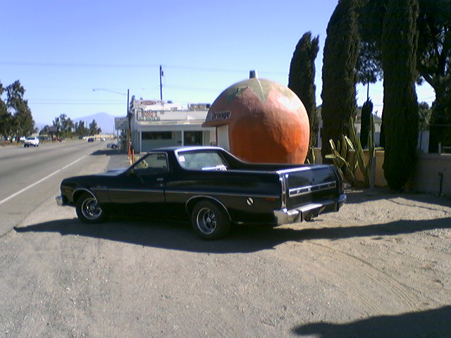 Black Ford Ranchero parked by giant orange-shaped roadside stand