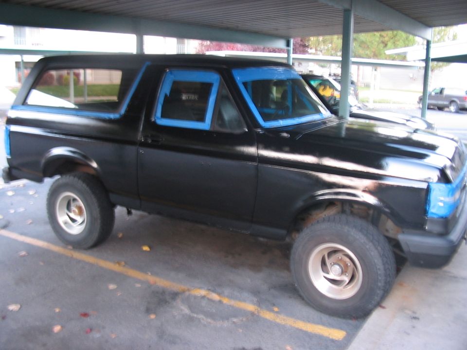 Black 1991 Ford Bronco parked under a carport, windows taped for painting